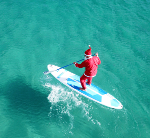 Als Weihnachtsmann verkleideter Mann paddelt auf einem Surfbrett im t&uuml;rkisblauen Meer in Australien.