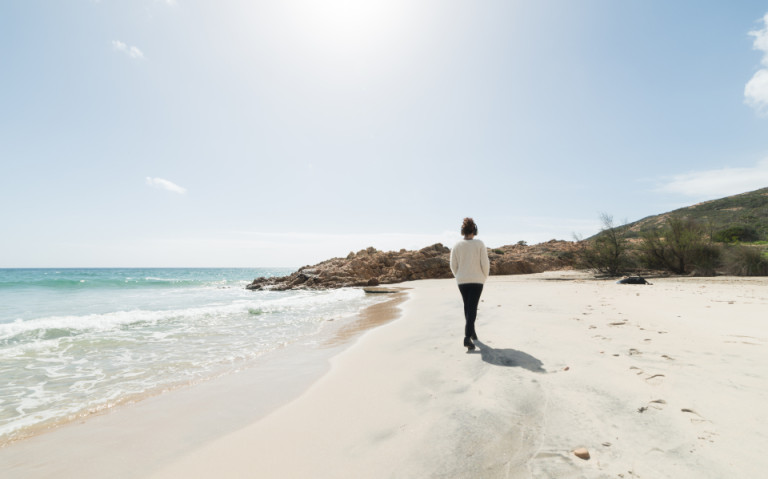 Eine Person spaziert am ruhigen Sandstrand entlang, während die Sonne scheint – eine Methode zur Anpassung des Tagesablaufs zur Vermeidung von Jetlag.