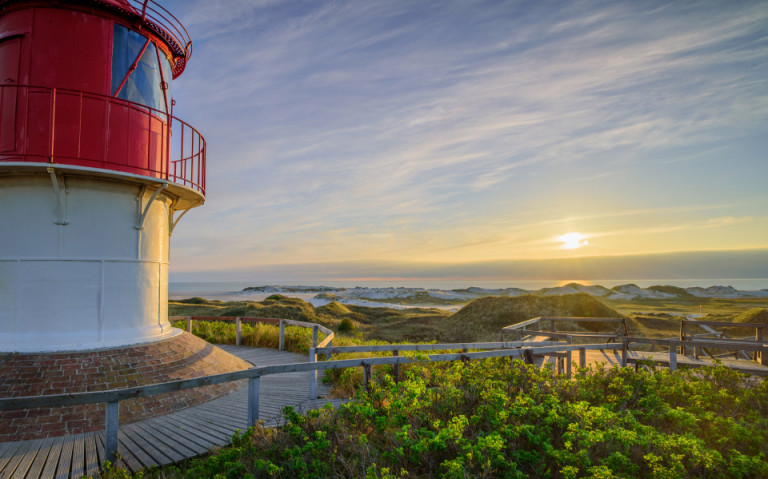 Der Leuchtturm auf der Insel Amrum, umgeben von grünen Dünen und dem weiten Blick auf die Nordsee bei Sonnenuntergang.
