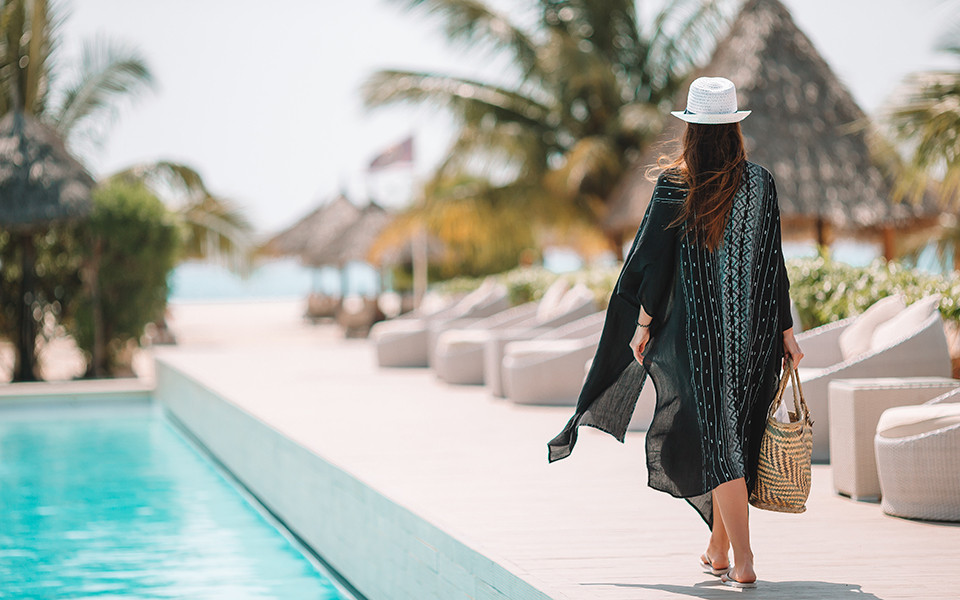 Frau im Sommerkleid mit Strohhut spaziert barfu&szlig; am Pool eines tropischen Resorts.