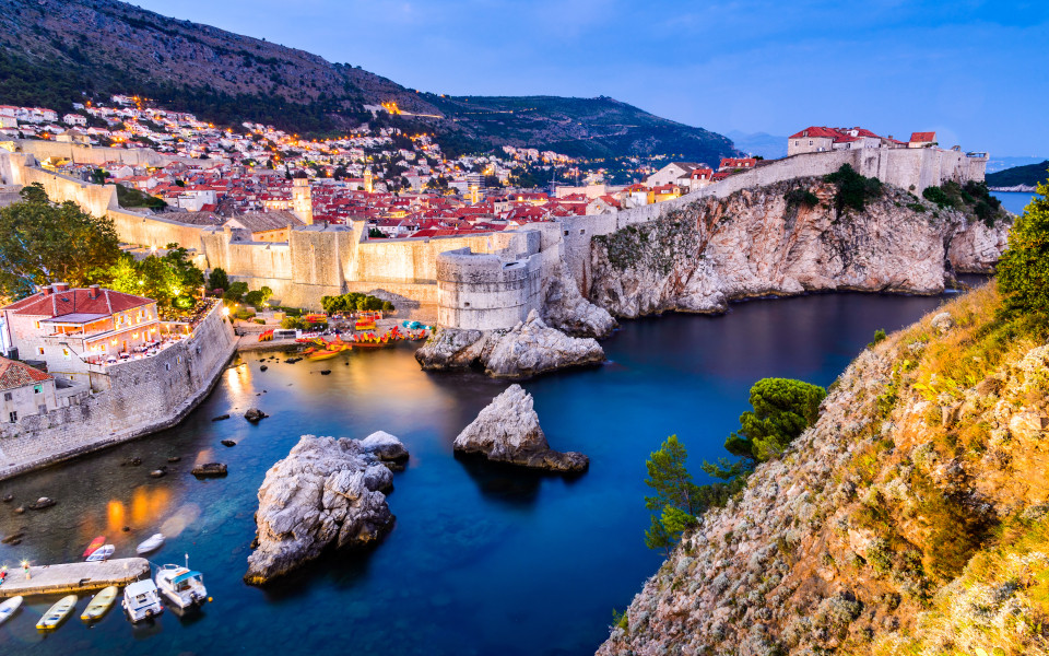 Blick auf die Altstadt von Dubrovnik mit ihren mittelalterlichen Stadtmauern und dem türkisblauen Wasser im Vordergrund.