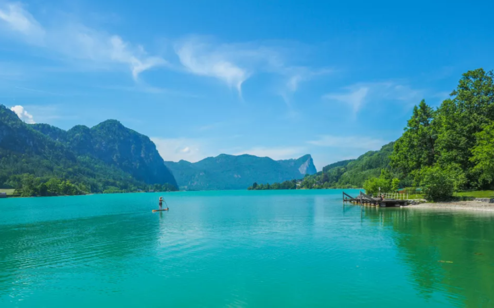 Stand-up-Paddler auf türkisblauem See im Salzkammergut, umgeben von bewaldeten Berghängen und Steg am rechten Ufer.