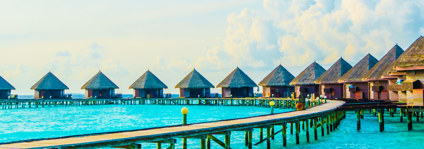 Reihen von Überwasser-Bungalows mit Strohdächern auf den Malediven, verbunden durch Holzstege über türkisblauem Meer, eingerahmt von Palmenblättern im Vordergrund.